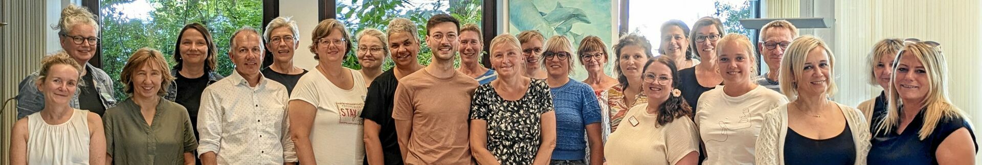 Das Bild zeigt 24 Personen in zwei Reihen. Es sind nur die Oberkörper abgebildet. Die Gruppe befindet sich in einem Raum. Durch die Fenster im Hintzergrund sind grüne Zweige erkennbar. Die Stimmung ist hell und freundlich.
